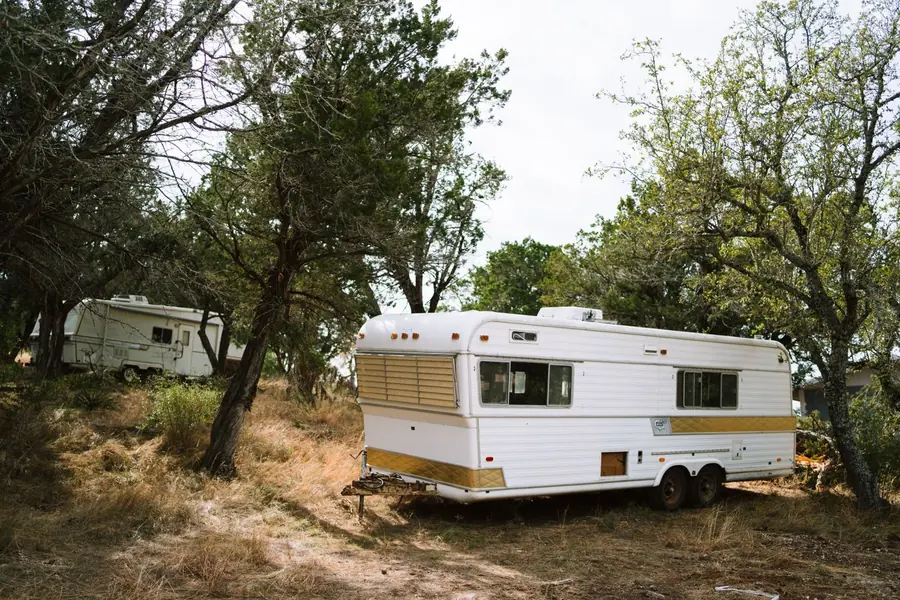 Older travel trailers parked among cedar and oak scrub on dry Texas ground